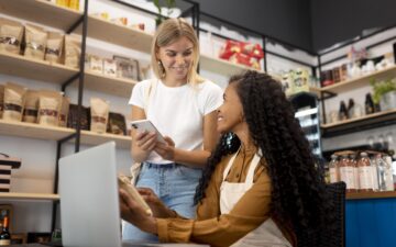 Duas mulheres sorridentes no caixa de uma loja física usando equipamentos digitais como notebook e celular.