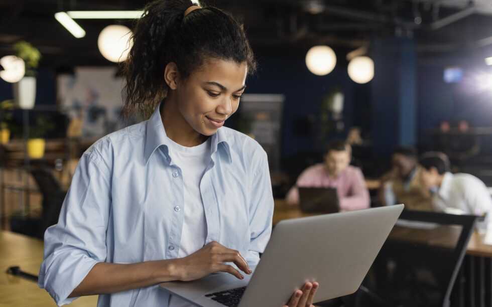 Uma jovem sorridente usando um notebook em um escritório moderno, com colegas ao fundo.