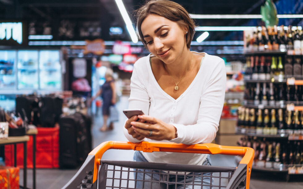 Mulher mexendo no celular enquanto caminha com o carrinho pelo supermercado.