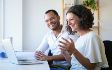 Duas pessoas sorrindo em uma videochamada pelo notebook, sentados à mesa.