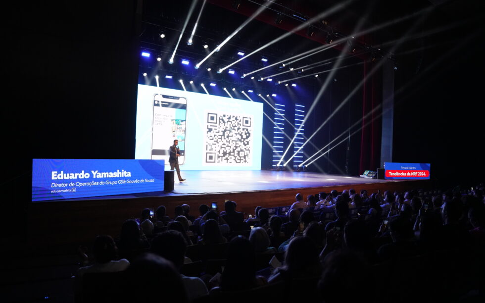 Imagem de um palestrante no palco do teatro na última edição do Cenários do Varejo. As luzes estão bem baixas, o palco super iluminado e, por trás do palestrante, há uma telão com QR Code.